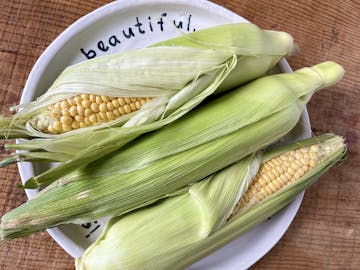 sweetcorn in it's husk on a white plate, laid on a wooden table