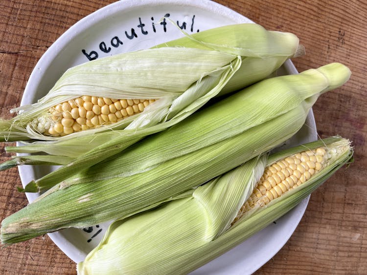 sweetcorn in it's husk on a white plate, laid on a wooden table
