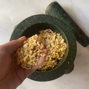 A handful of crushed breadcrumbs with hazelnuts and fennel seeds above a mortar and pestle. 
