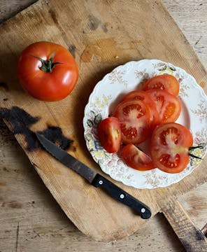 plate of sliced tomato, half a tomato on the side with knife and a whole tomato on top of a wooden board