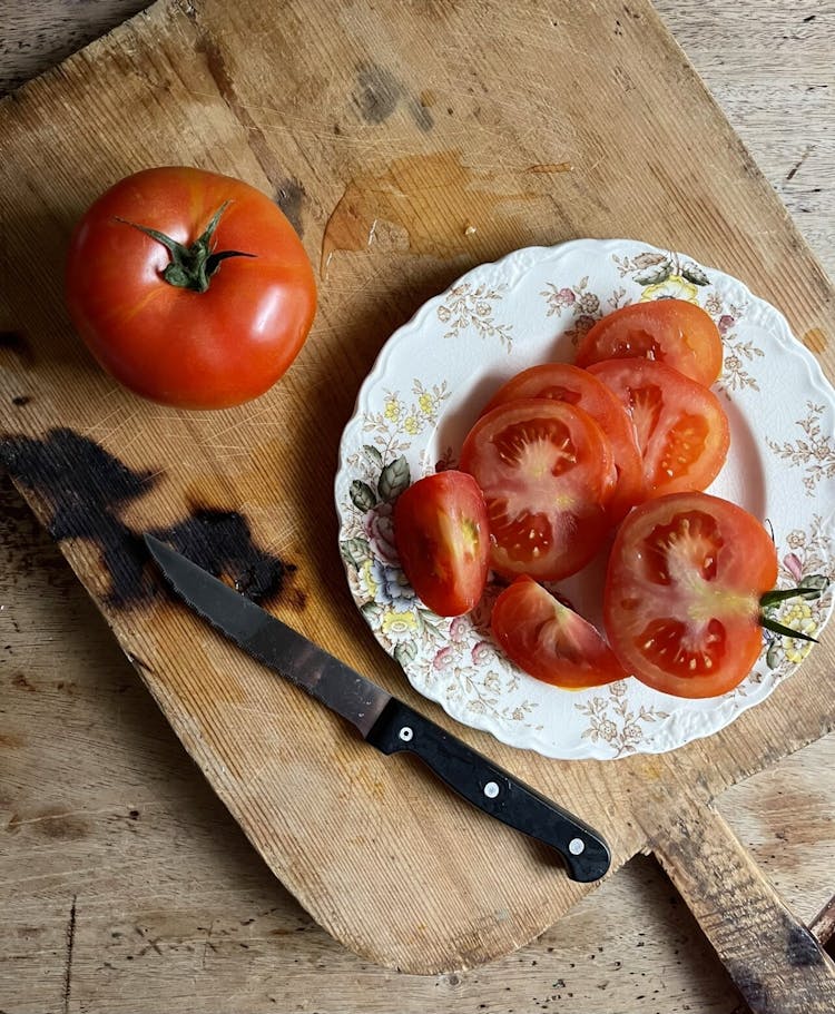 plate of sliced tomato, half a tomato on the side with knife and a whole tomato on top of a wooden board 