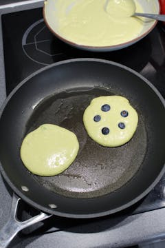 frying pan with 2 pancakes 1 topped with 5 bluberries