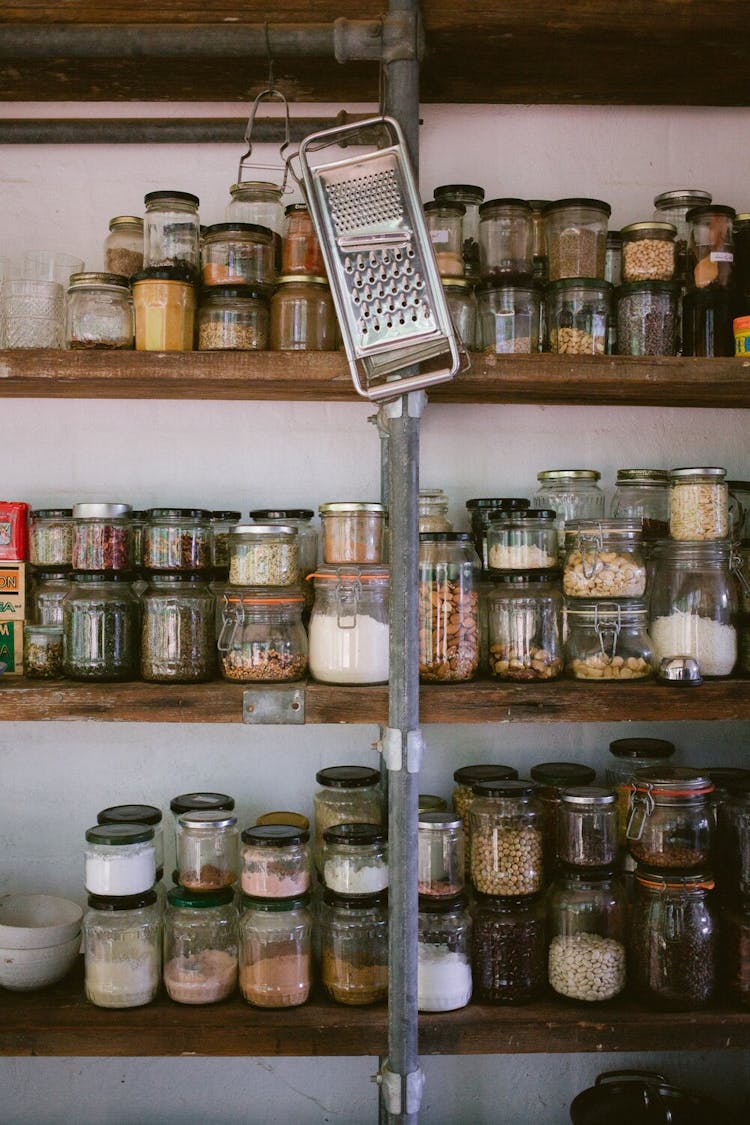 A grater hanging in front of 3 shelves full of jarred spices and other dried goods.