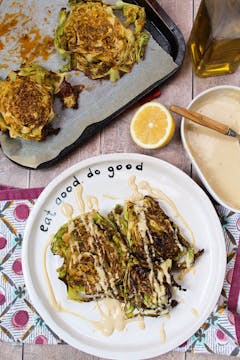 2 slices of roasted savoy cabbage steak drizzled with tahini dressing on white plate.half a lemon on the side and tahini dressing on a serving bowl. 2 more roasted savoy cabbage steaks on a baking tray