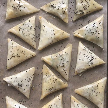 Raw unbaked samosas on a tray covered in onion seeds.