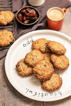 A plate of date, tahini and oat cookies. There's a cup of coffee, a bowl of dates, and a drying rack with more cookies on the table as well.