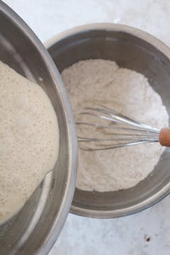 Pouring a milk and cream mixture into a bowl of flour, a whisk is visible in the bowl of flour.