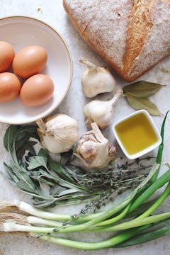 bunch of spring onion, thyme, 4 whole garlic, 2 bay leaves, little bowl of oil, 4 whole eggs on a bowl and bread