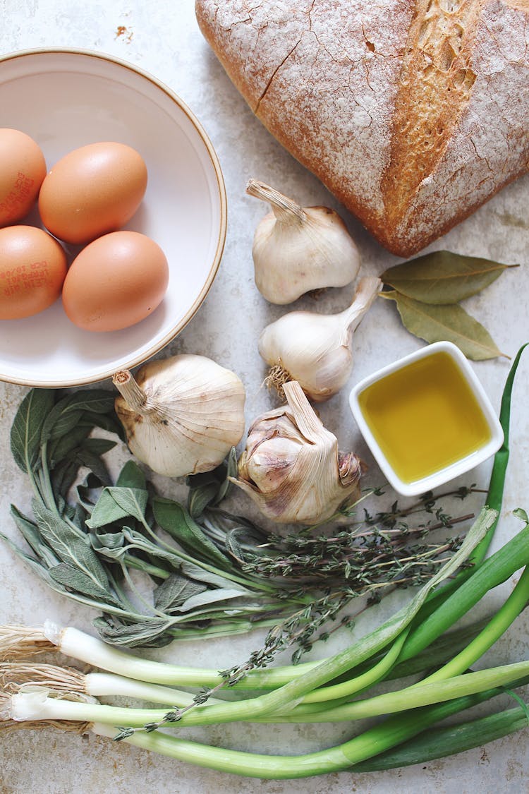 bunch of spring onion, thyme, 4 whole garlic, 2 bay leaves, little bowl of oil, 4 whole eggs on a bowl and bread