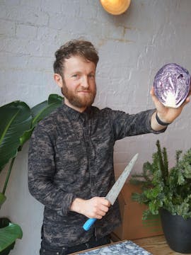 Douglas McMaster holding a purple cabbage and a knife, about to prep it. 