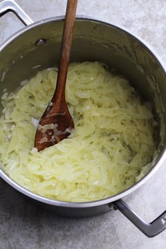 large saucepan with sliced oinion being cooked with olive oil, wooden spoon is being used to stir 