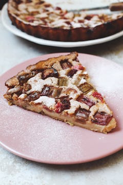 A slice of vegan rhubarb clafoutis on a pink plate, dusted with icing sugar. The remaining half of the clafloutis is visible just behind this plate.