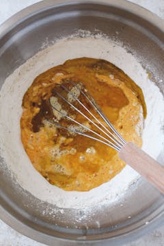 A large mixing bowl with flour, herbes de provence, salt, and pepper. There is a well in the centre where eggs, olive oil, mustard, and milk have been added. There's a whisk mixing everything together. 