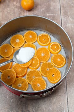 Thinly sliced clementines at the bottom of a cake tin. There's a spoonful of sugar above the tin, about to be sprinkled on top. 