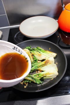 Pak choi cut in half, frying in a pan. The soy sauce mixture is being poured to deglaze. 