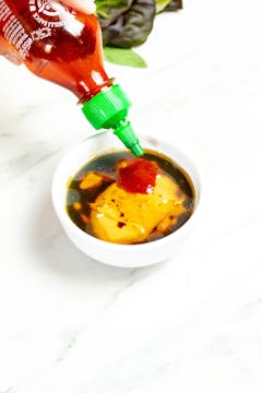 Sriracha being added to the ingredients for the peanut dipping sauce in a small bowl. Already in the bowl is: peanut butter, lime juice, soy sauce, and sesame oil.