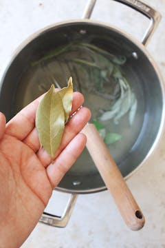 A hand holding 2 bay leaves above a pot of herbs. 