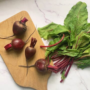 4 beetroots on a chopping board. Their stems have been separated, and are next to the board with their leaves attached.
