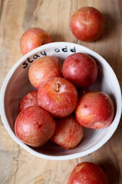 A bowl of plums. There are a couple scattered on the countertop below it. 