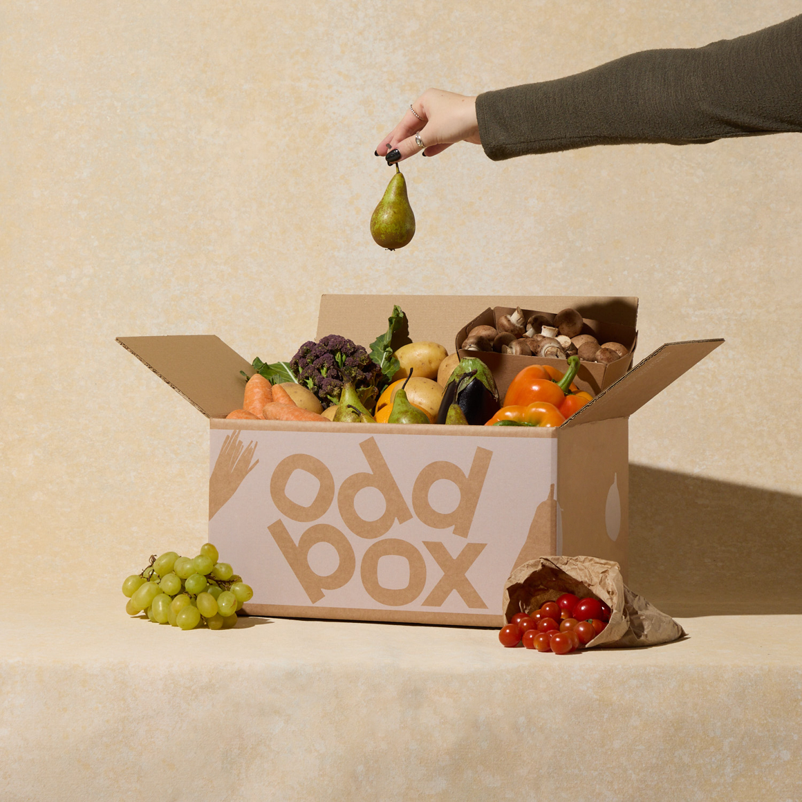 Oddbox filled with fruit and veg and a hand holding a pear.