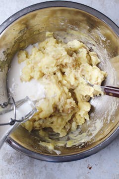 In a mixing bowl, mashed potato and milk getting poured onto it 