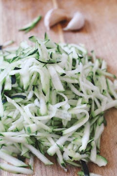 A pile of grated courgette on a chopping board. 2 garlic cloves are visible in the background. 