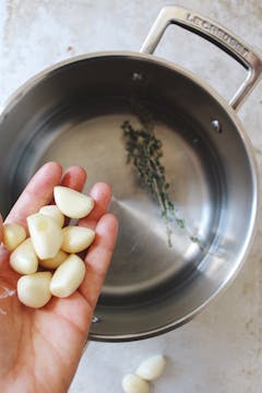 A hand holding 9 pieces of peeled garlic above a bit with sprigs of herbs in it.