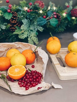 Oranges wrapped in baking paper with cranberries and herbs. There's a chopping board with 4 more oranges on top; a Christmas table wreath is visible beyond the oranges.