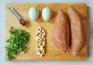 A chopping board with all the ingredients you need for sweet potato baked eggs. Eggs, sweet potatoes, chilli flakes, coriander, and cashews. 