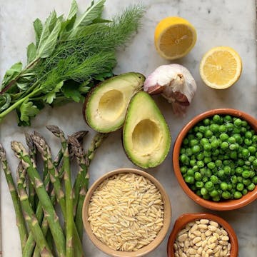 All the ingredients required for avocado spring pasta, including orzo, pine nuts, garlic cloves, peas, asparagus, herbs, avocados, and two halves of a lemon.