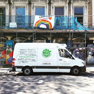 A white van with the City Harvest logo on it. There is scaffolding on the building behind the van, with a sheet with a rainbow on it dedicated to the NHS hanging off of it.