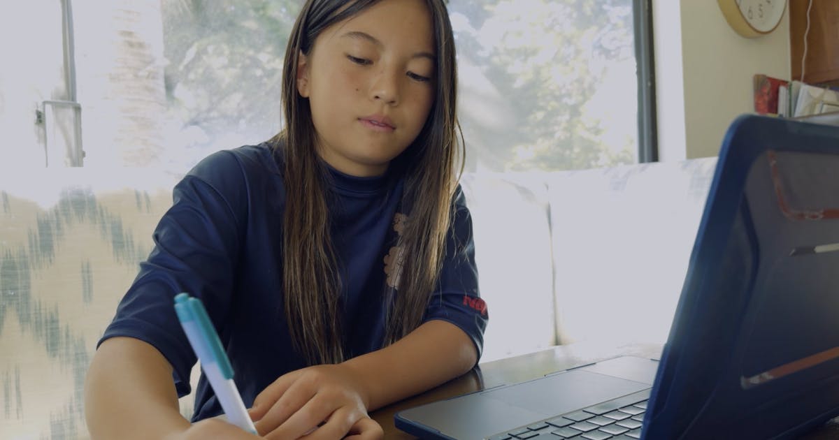 Photo of a child working on their laptop and taking handwritten notes.