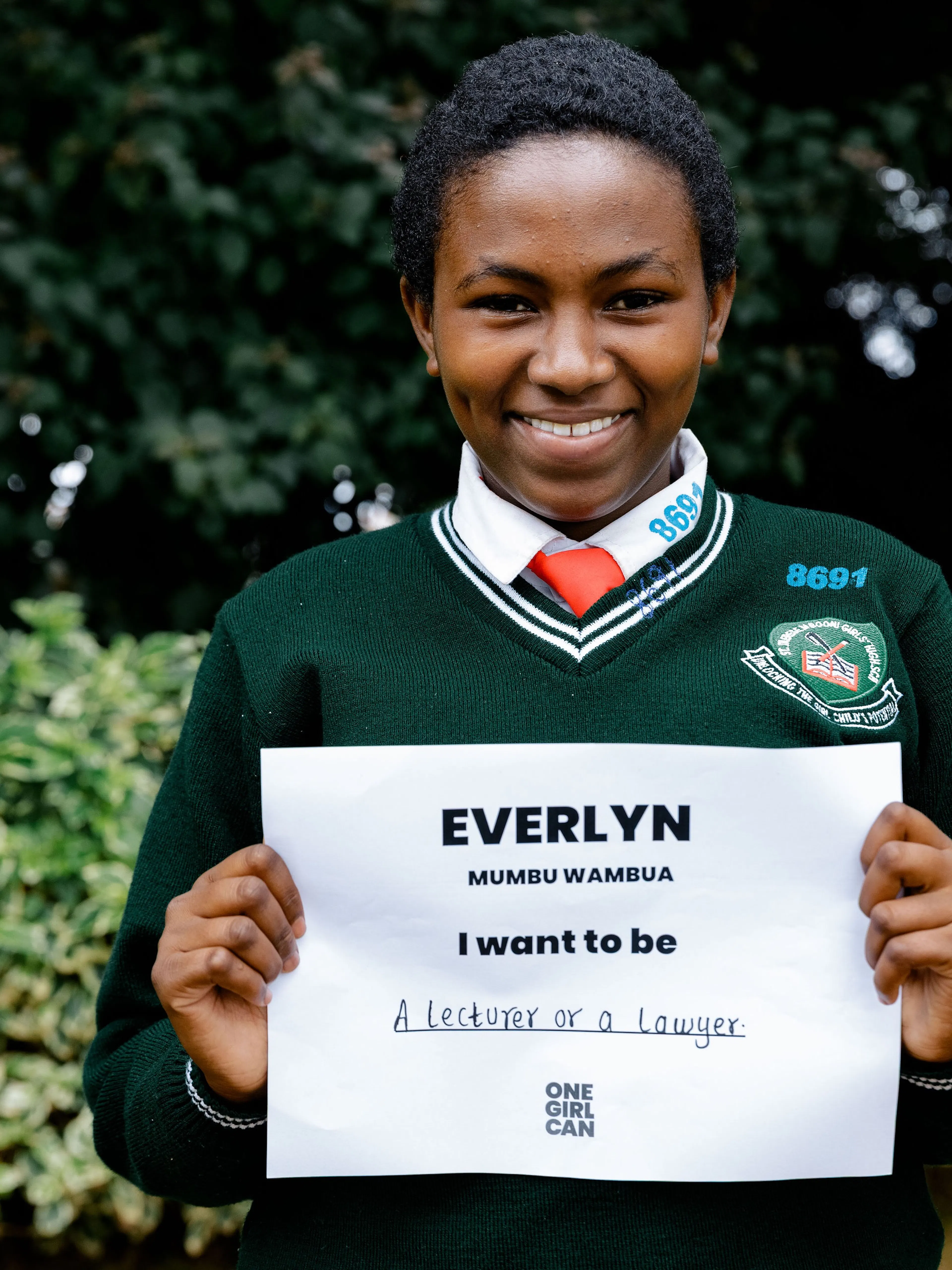 Everlyn, a One Girl Can scholarship candidate in school uniform, holding a handwritten sign that reads “I want to be a Lecturer or Lawyer”