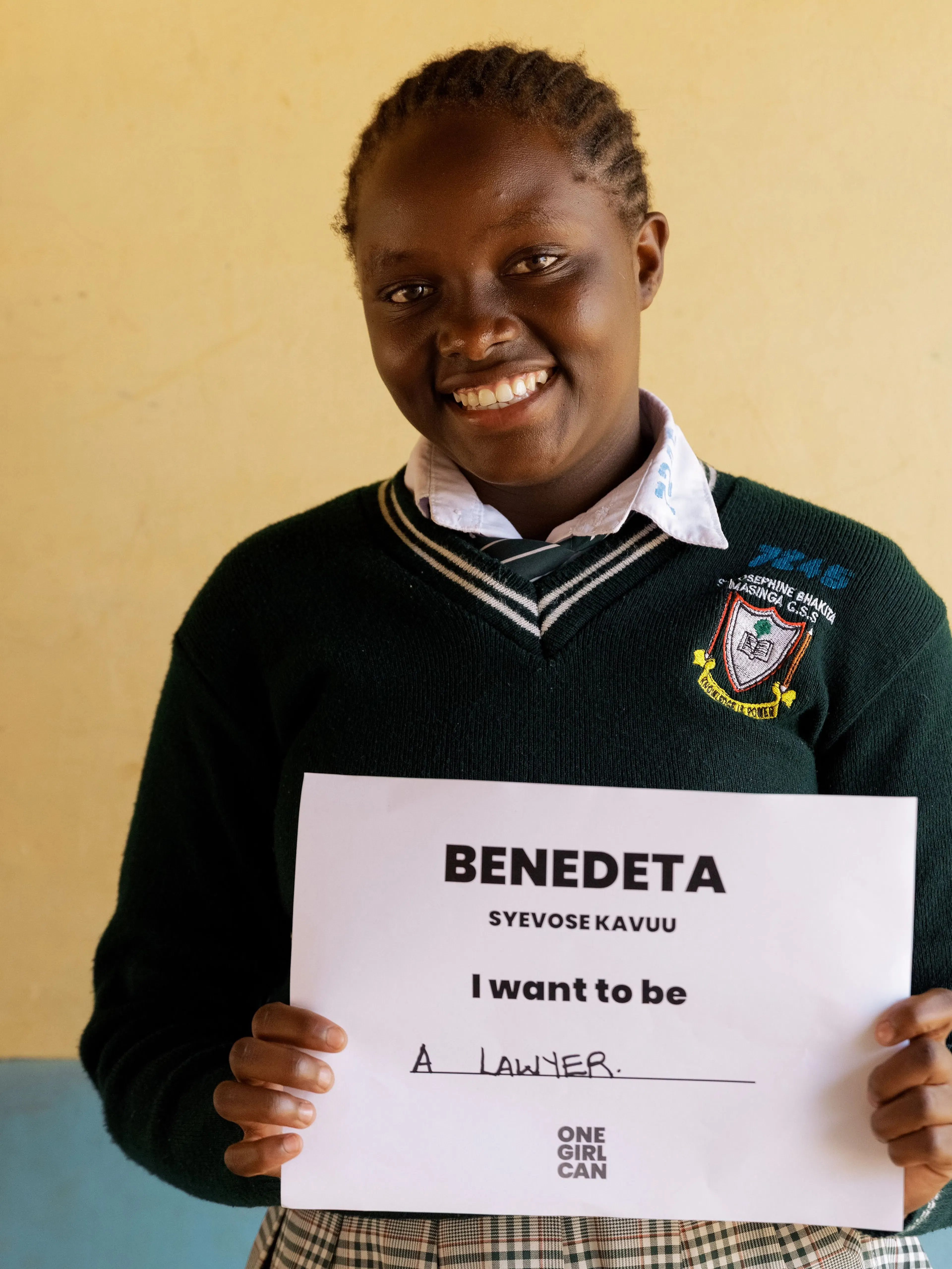 Benedeta, a One Girl Can scholarship candidate in school uniform, holding a handwritten sign that reads “I want to be a Lawyer”