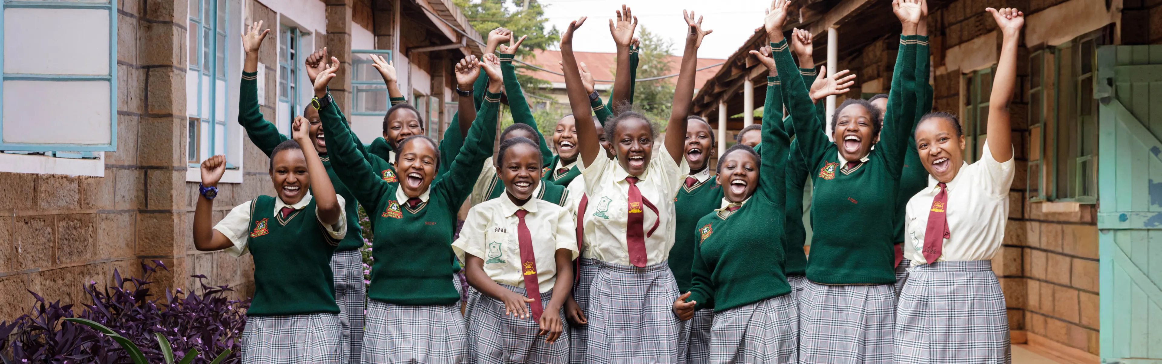 A group of students in green school uniforms cheering with their arms raised outside their school in Kenya