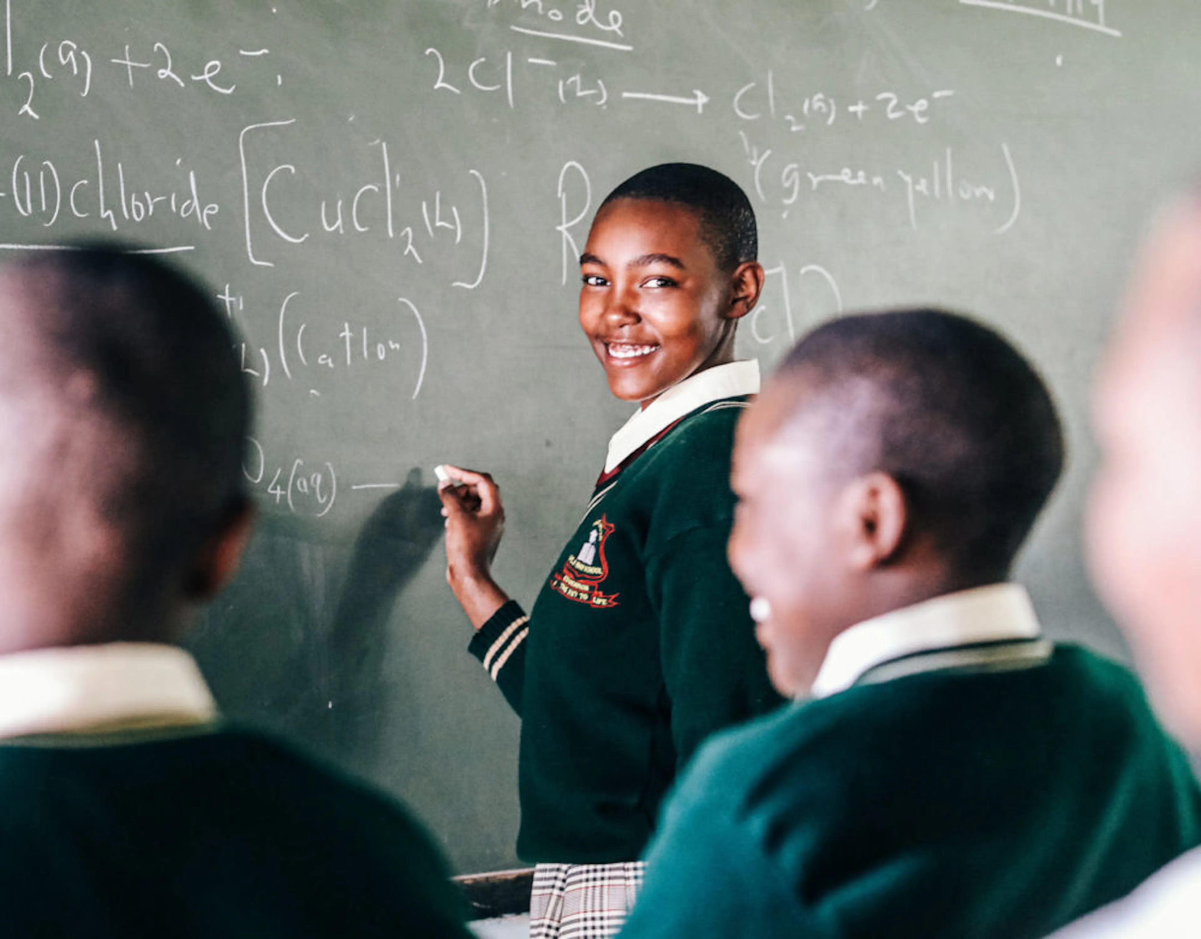Student smiling as she writes a chemistry equation on a chalkboard in front of her class
