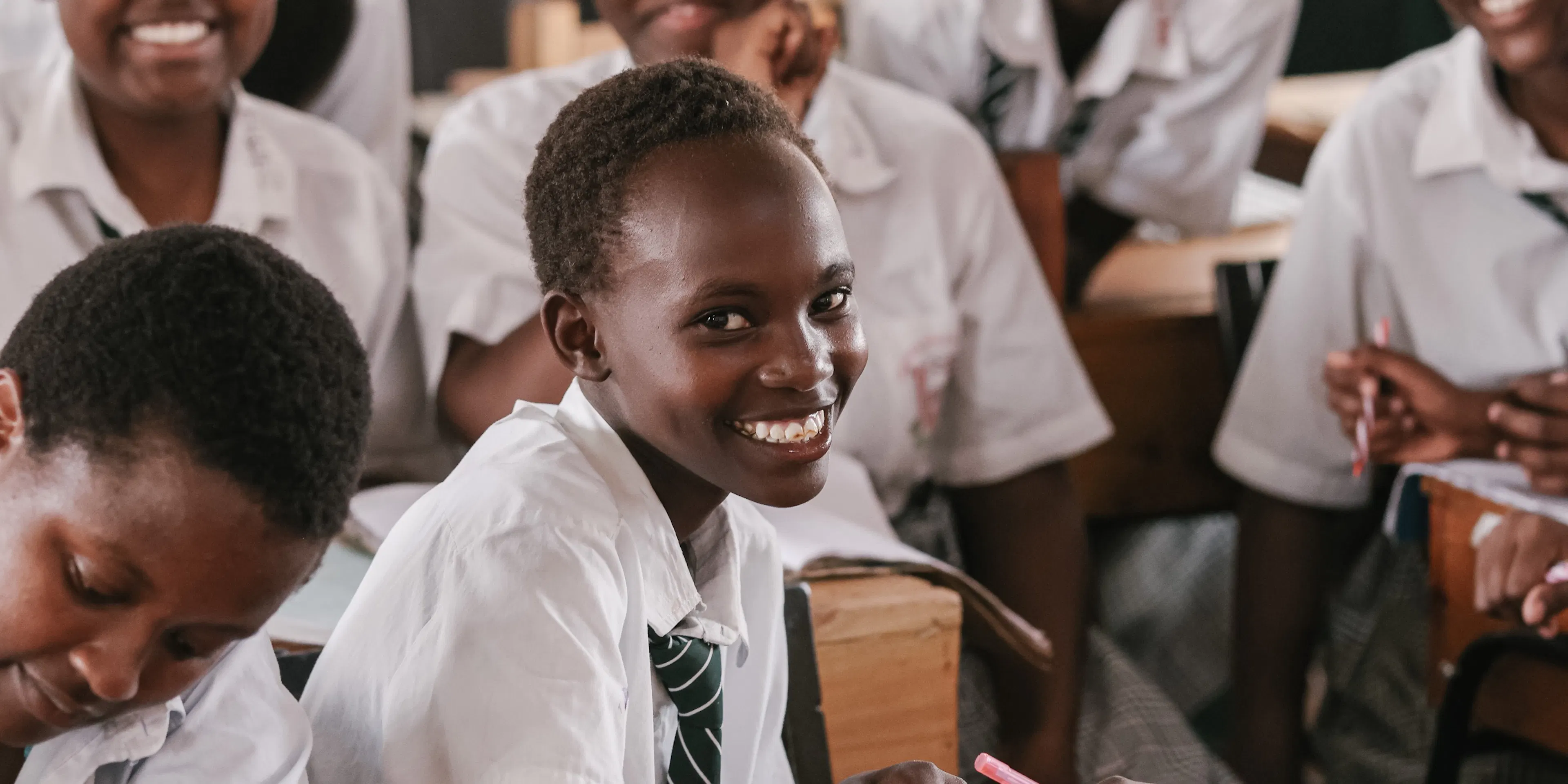 A student in a white uniform shirt smiling over her shoulder in a busy classroom
