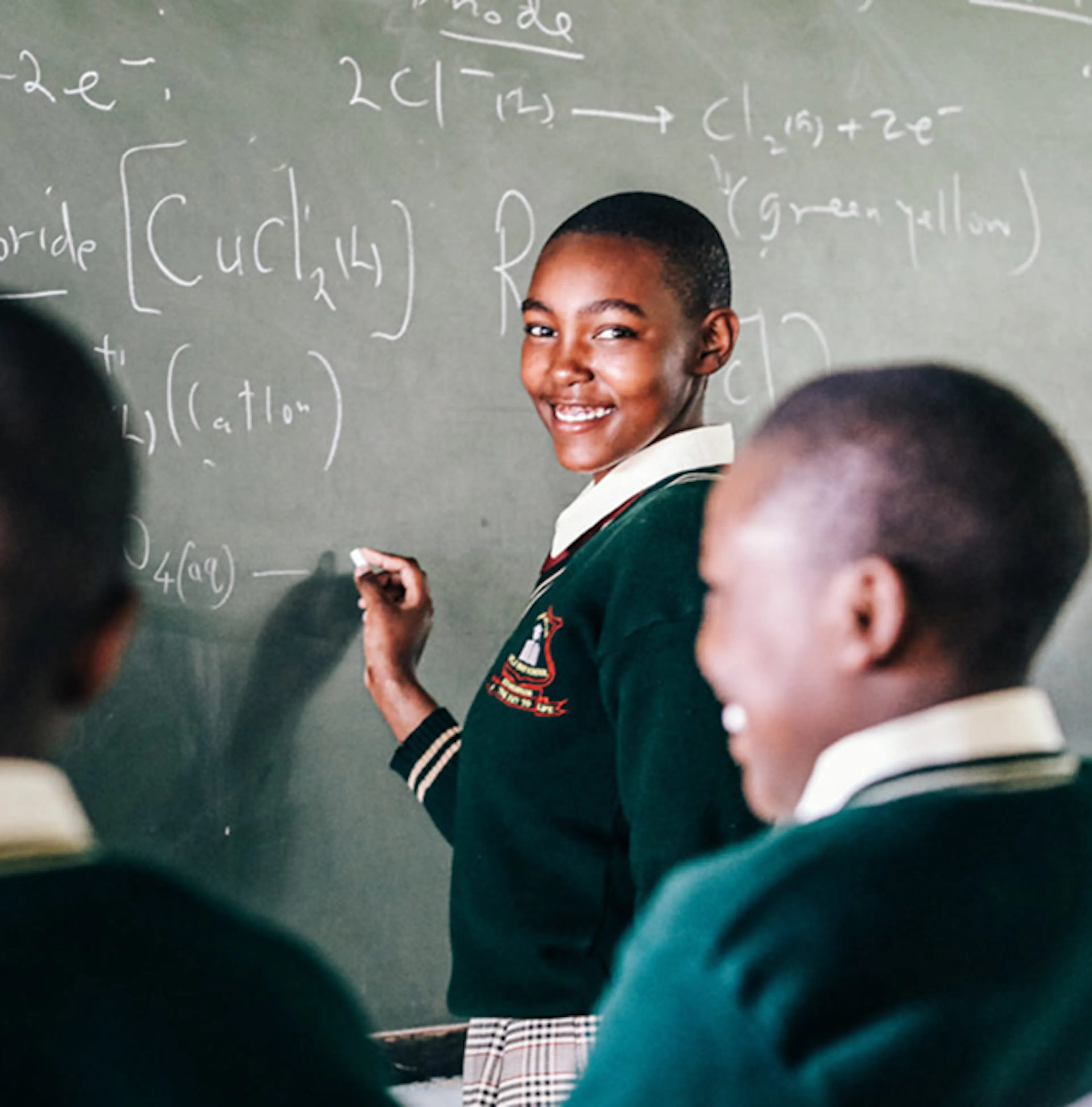 A student in a green school uniform smiling at the camera while writing a chemistry equation on a chalkboard