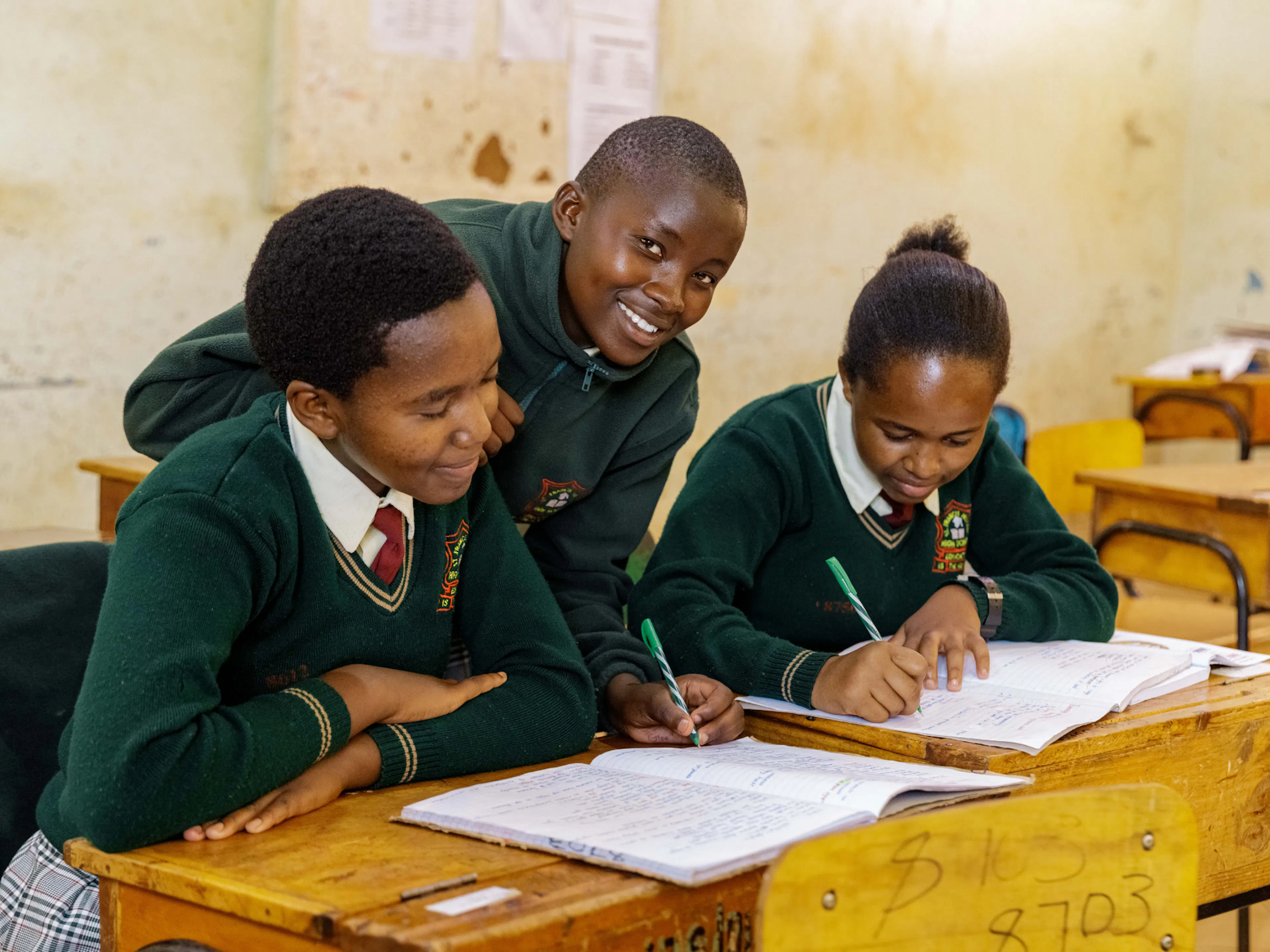 Three students in green uniforms working together on schoolwork at their desks