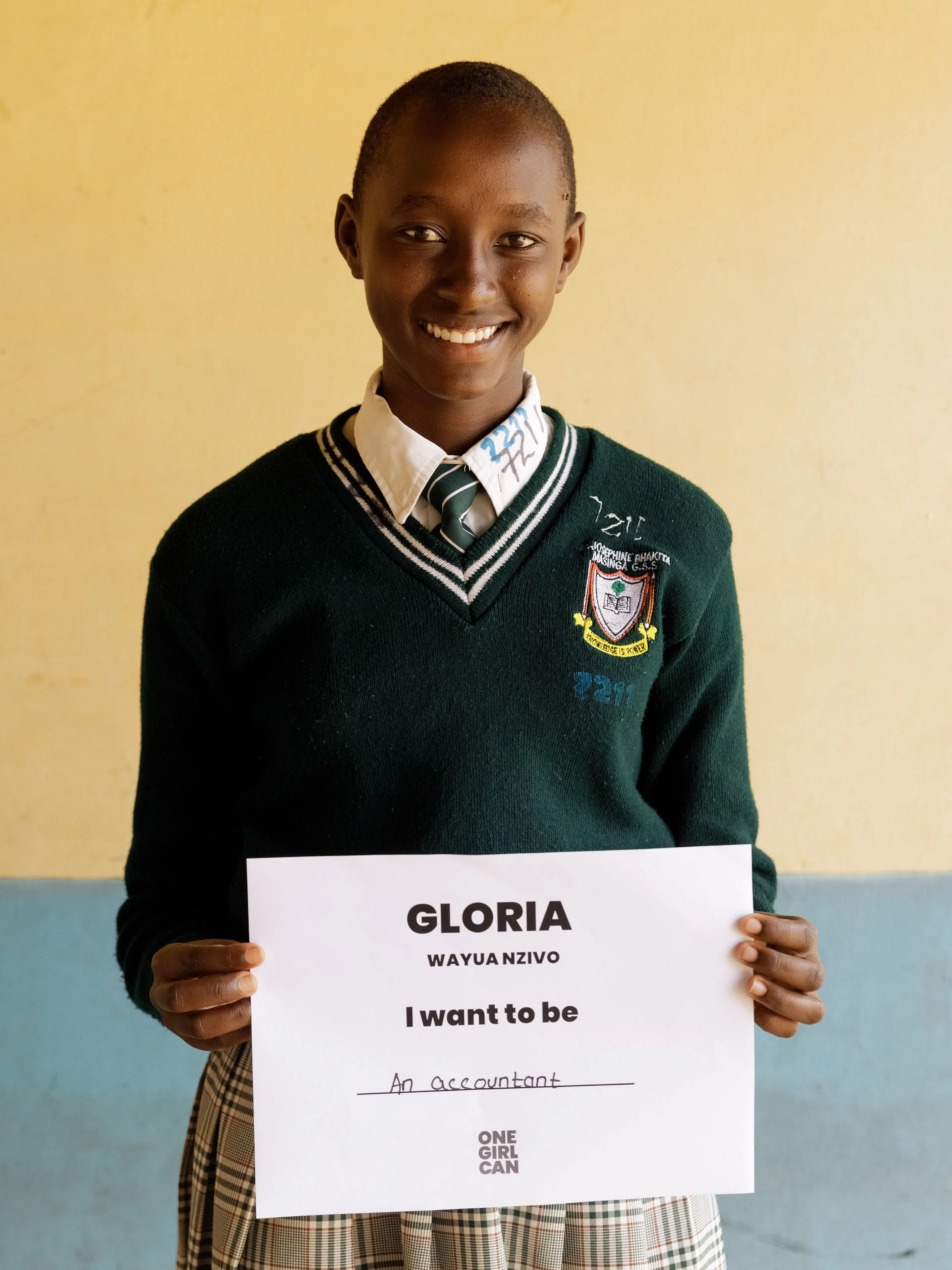 Gloria, a One Girl Can scholarship candidate in school uniform, holding a handwritten sign that reads “I want to be an Accountant”