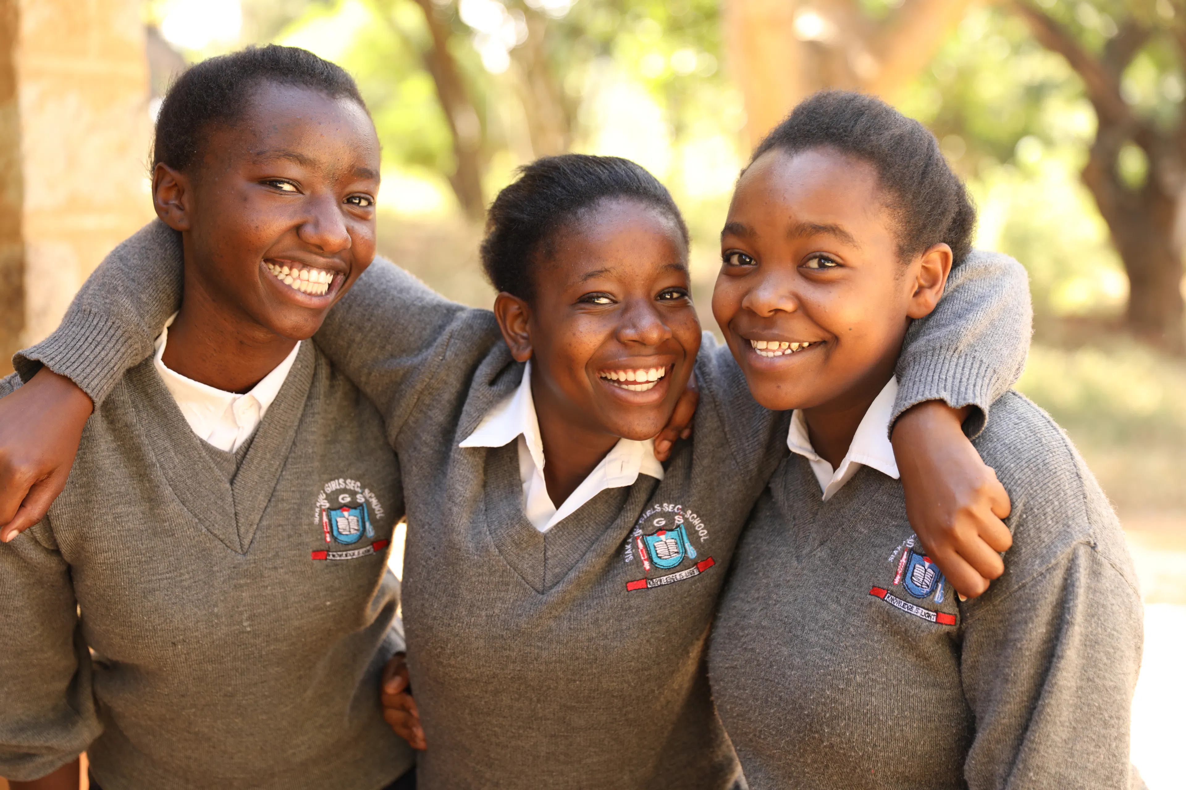 Three secondary school students in grey uniforms with their arms around each other, smiling