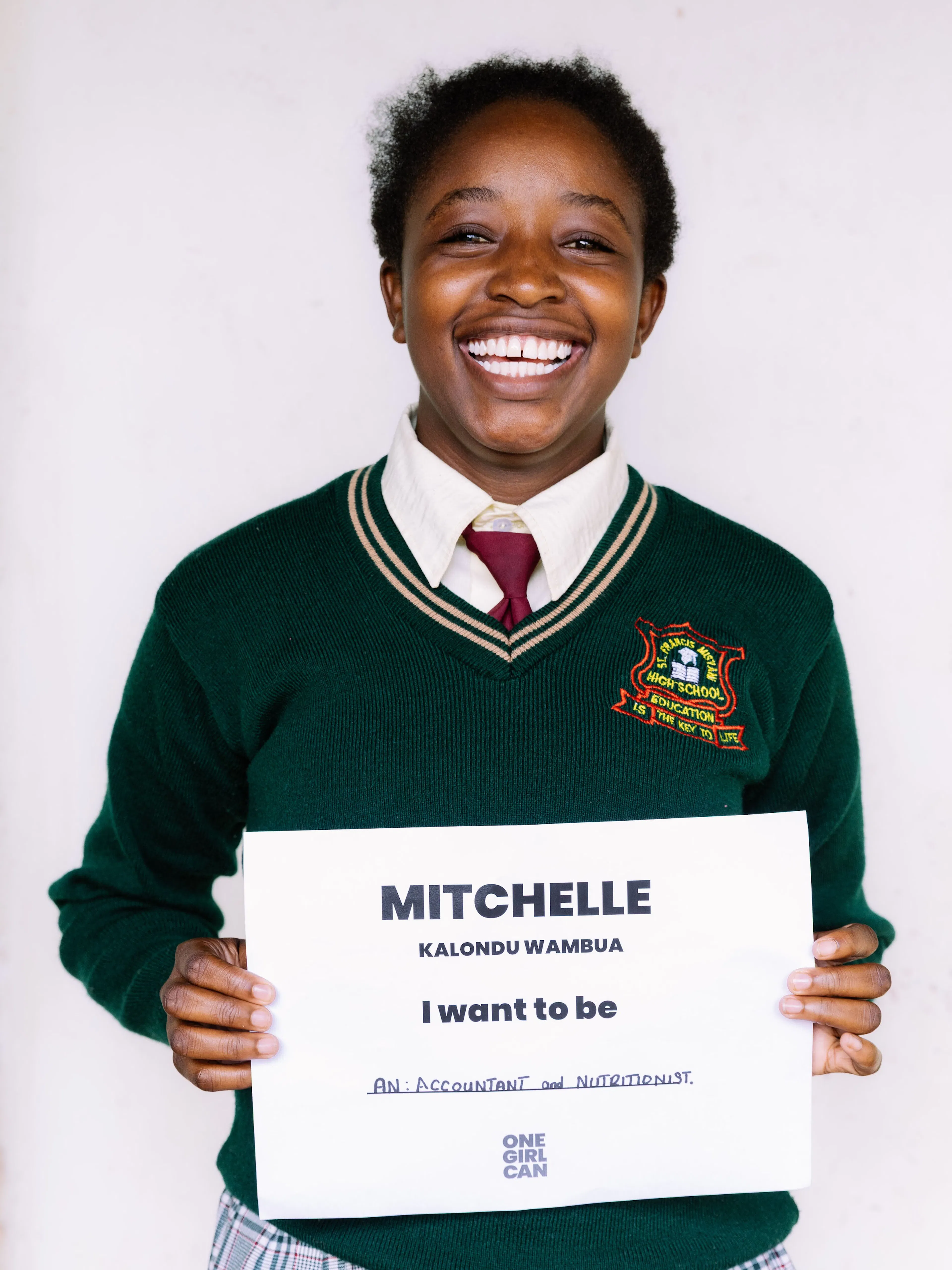 Mitchelle, a One Girl Can scholarship candidate in school uniform, holding a handwritten sign that reads “I want to be an Accountant or Nutritionist”