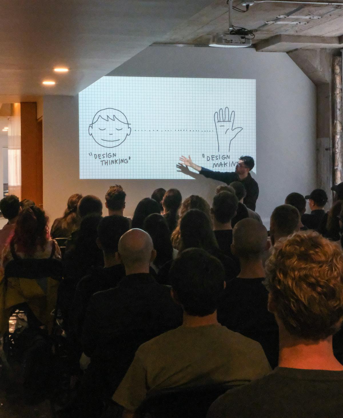 A man with glasses standing in front of a wall gestures towards a projection of a drawing that says 'design thinking...design making' with his right arm, in front of a crowd of people sitting in chairs, facing the man and the projection.
