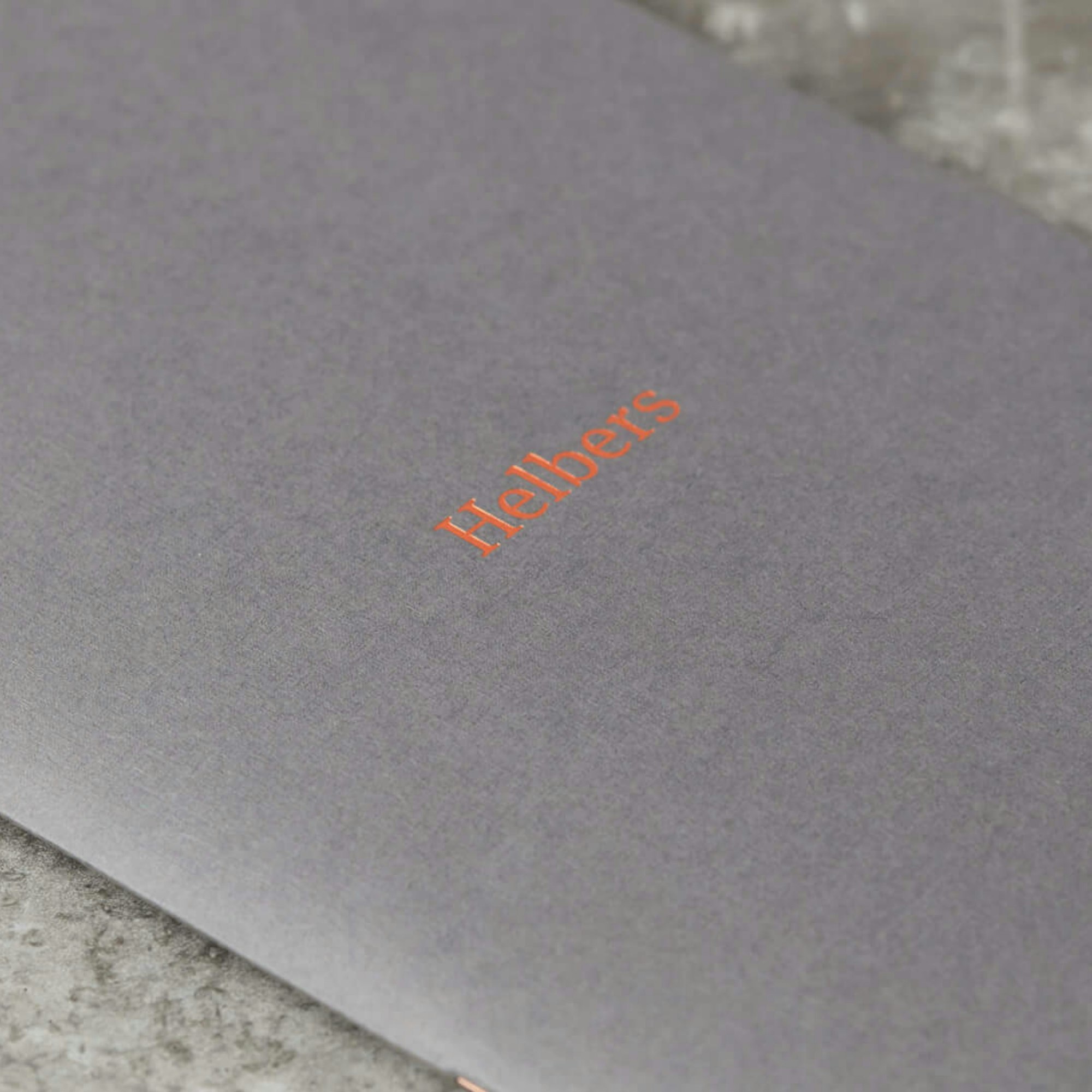 A print of the Helbers menswear brochure, with a grey cover and an embossed Helbers logotype in orange.