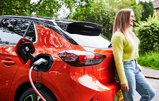 Woman leaning against an orange Corsa e, with an electric car charger plugged into it