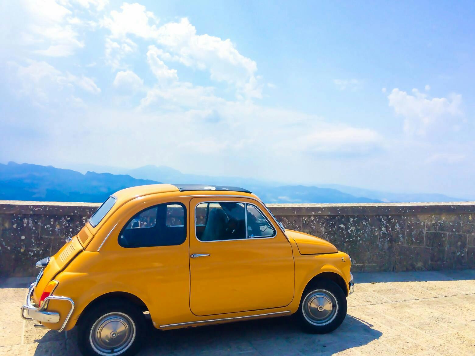 A yellow Fiat 500 is park in front of a low wall overlooking a wide vista with blue sky.