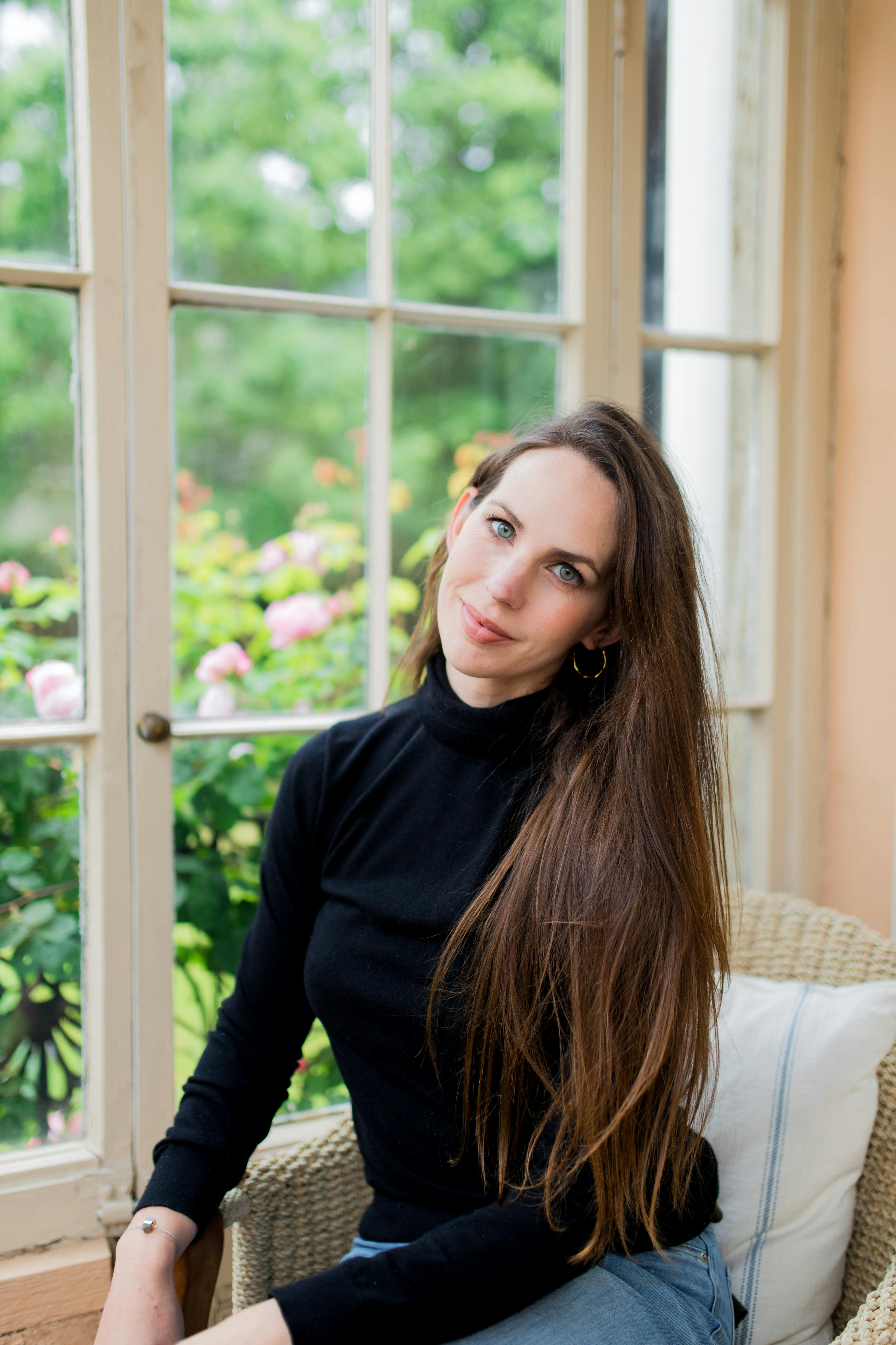 A photograph of Hayley Quinn sitting in a chair facing towards the camera and smiling with her head tilted slightly on one side. In the background is a window looking out into a green garden with a rosebush close to the window.