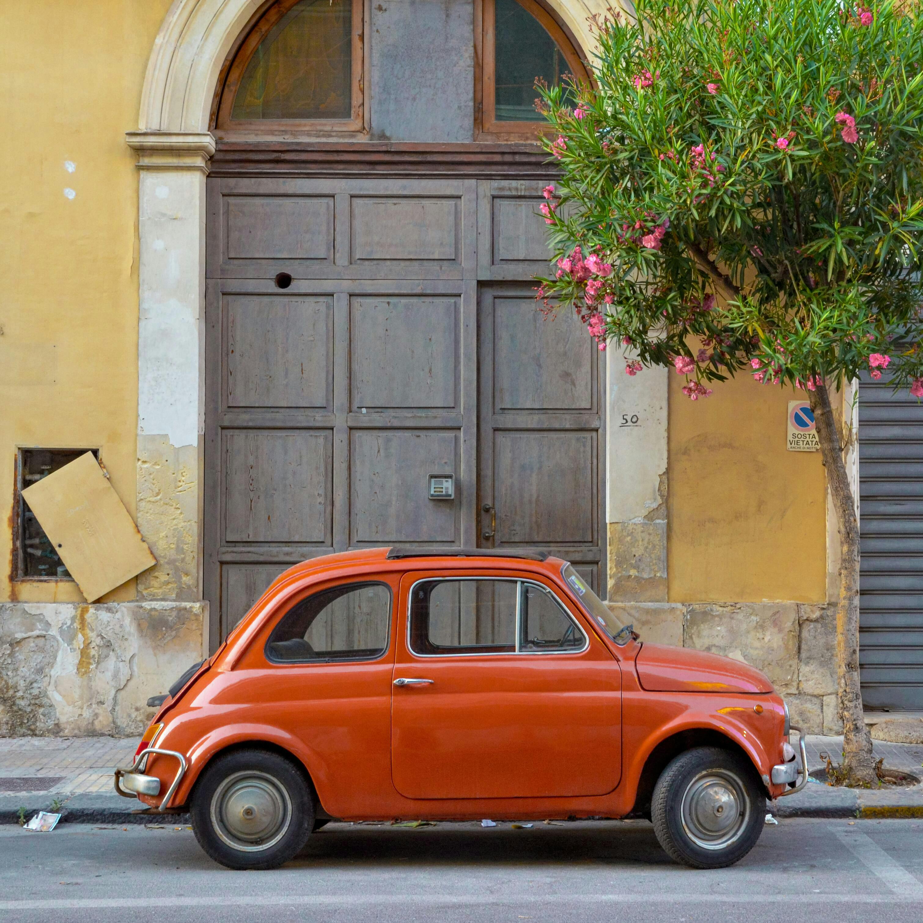 An orange Fiat 500 is parked side-on on an Italian street in front of large wooden doors. On the right hand side is a tree in bloom.