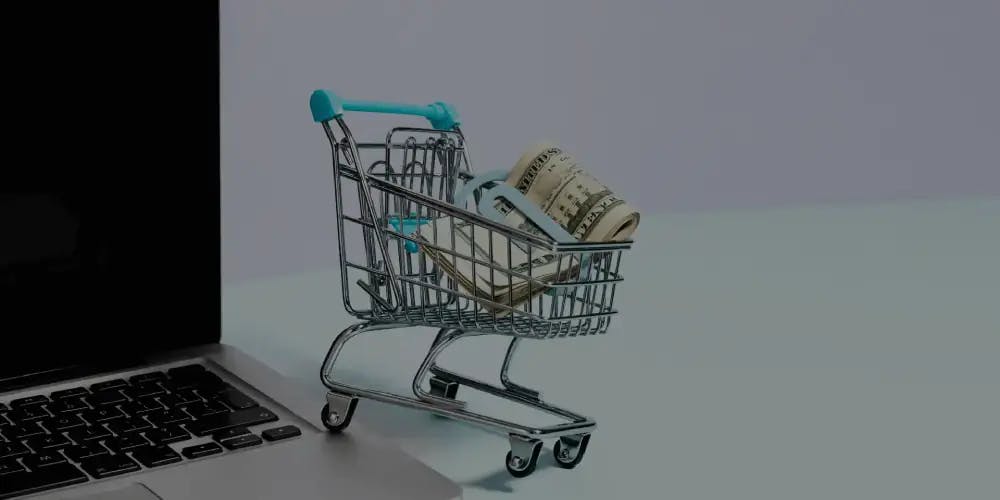 Miniature shopping card with bankroll in it, next to a laptop.