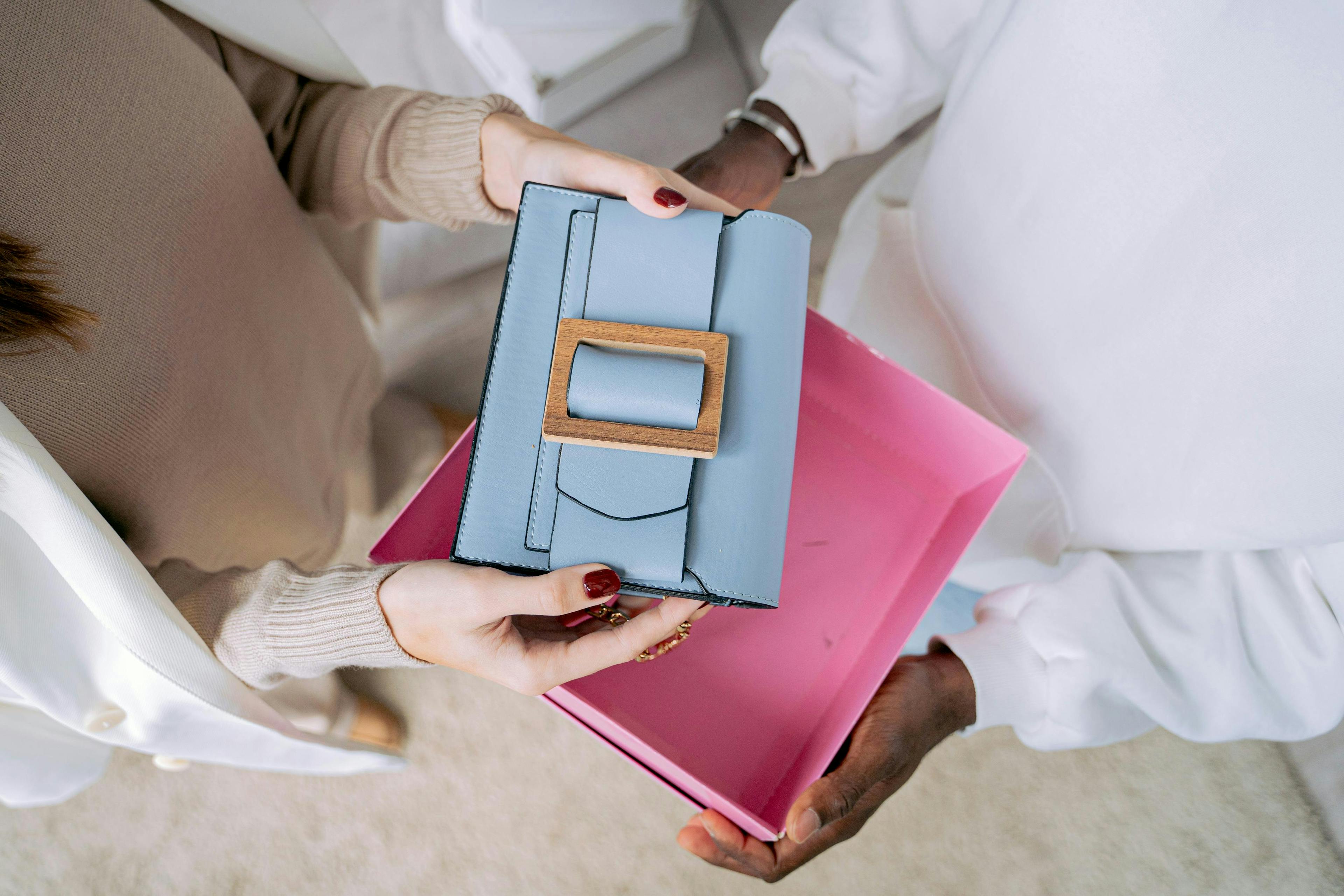 Hands exchanging a light blue wallet with wooden buckle detail over a pink surface
